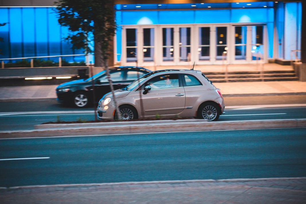 Fiat 500 jedzie nocą obok nowoczesnego, niebiesko podświetlonego budynku Fiat 500 jedzie nocą obok nowoczesnego, niebiesko podświetlonego budynku